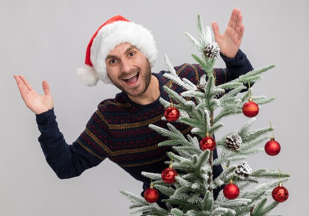 Impresionado joven caucásico con sombrero de navidad de pie detrás del árbol de navidad mirando a la cámara manteniendo las manos en el aire aislado sobre fondo blanco.