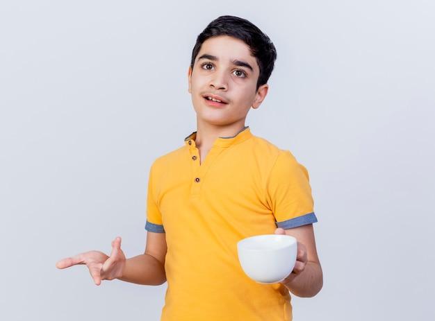 Impresionado joven caucásico sosteniendo la taza mirando y apuntando directamente aislado sobre fondo blanco con espacio de copia