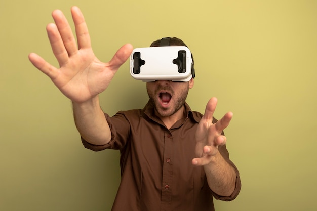 Impresionado joven caucásico vistiendo auriculares vr estirando las manos hacia la cámara mirando al lado con la boca abierta aislada sobre fondo verde oliva con espacio de copia