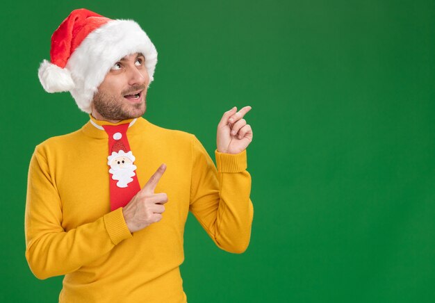 Impresionado joven caucásico vistiendo gorro de navidad y corbata mirando y apuntando hacia arriba aislado sobre fondo verde