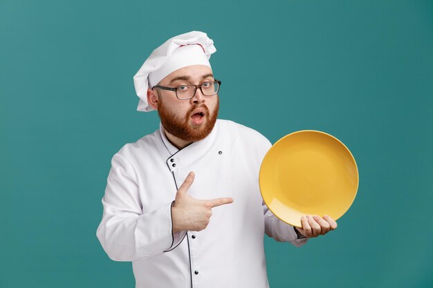 Impresionado joven chef masculino con uniforme de anteojos y gorra mostrando y señalando un plato vacío mirando a la cámara aislada en el fondo azul