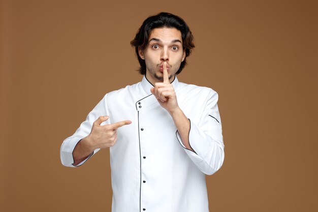 Impresionado joven chef masculino vistiendo uniforme mostrando gesto de silencio mirando a la cámara apuntándose a sí mismo aislado sobre fondo marrón