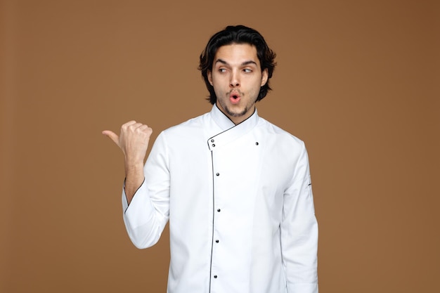 Impresionado joven chef uniformado mirando al lado apuntando al lado aislado de fondo marrón