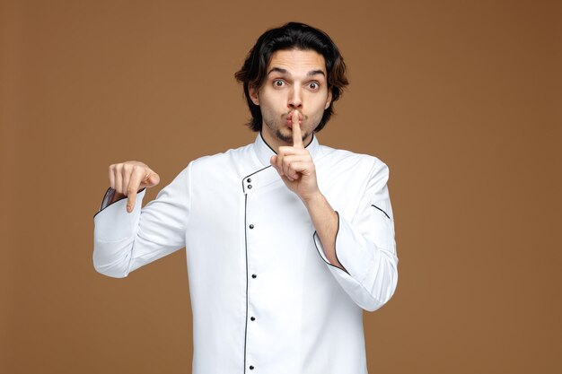 Impresionado joven chef uniformado mirando a la cámara mostrando un gesto de silencio apuntando hacia abajo aislado en un fondo marrón