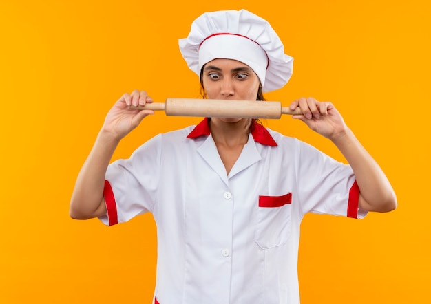 Impresionado joven cocinera vistiendo uniforme de chef sosteniendo y mirando el rodillo en la pared amarilla aislada