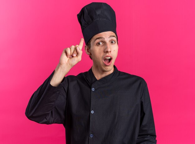Impresionado joven cocinero rubio en uniforme de chef y gorra mirando a la cámara apuntando hacia arriba aislado en la pared rosa
