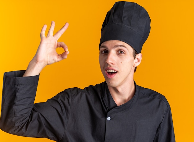 Impresionado joven cocinero de sexo masculino rubio en uniforme de chef y gorra mirando a la cámara haciendo bien firmar aislado en la pared naranja