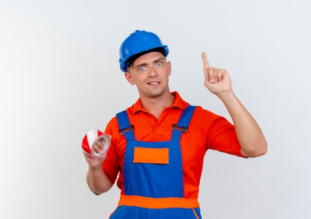 Impresionado joven constructor con uniforme y casco de seguridad sosteniendo cinta adhesiva y apunta hacia arriba
