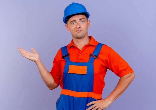 Impresionado joven constructor vistiendo uniformes y puntos de casco de seguridad con la mano al lado