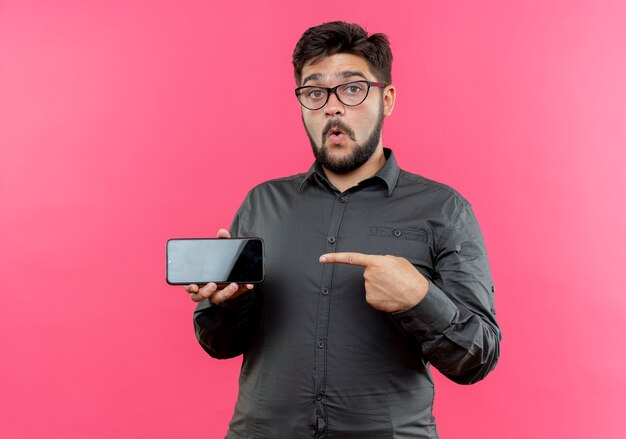 Impresionado joven empresario con gafas sosteniendo y apunta al teléfono aislado en la pared rosa