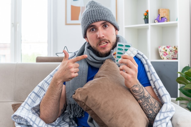 Foto gratuita impresionado joven enfermo con bufanda y gorro de invierno envuelto en una manta sentado en el sofá en la sala de estar sosteniendo una almohada y paquetes de píldoras mirando a la cámara apuntando a las píldoras