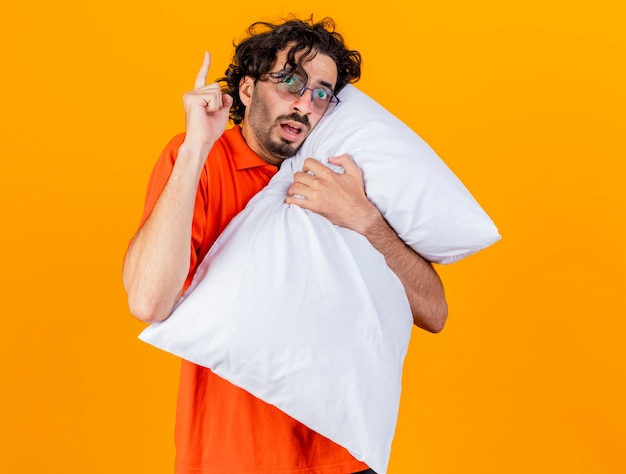 Impresionado joven enfermo caucásico con gafas abrazando la almohada mirando a la cámara levantando el dedo aislado sobre fondo naranja con espacio de copia