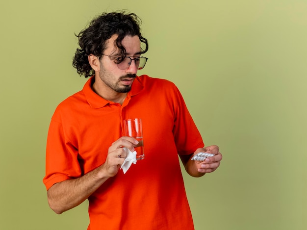 Impresionado joven enfermo con gafas sosteniendo un vaso de agua paquete de tabletas y una servilleta mirando tabletas aisladas en la pared verde oliva con espacio de copia