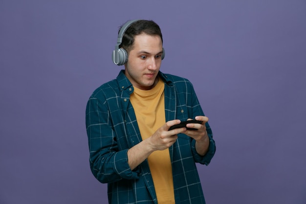 Impresionado joven estudiante masculino usando auriculares jugando en el teléfono móvil aislado sobre fondo púrpura