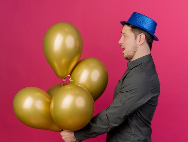 Impresionado joven fiestero con sombrero azul sosteniendo y mirando globos aislados en rosa