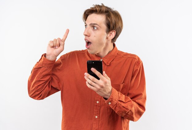 Impresionado joven guapo con camisa roja sosteniendo puntos de teléfono en arriba aislado en la pared blanca