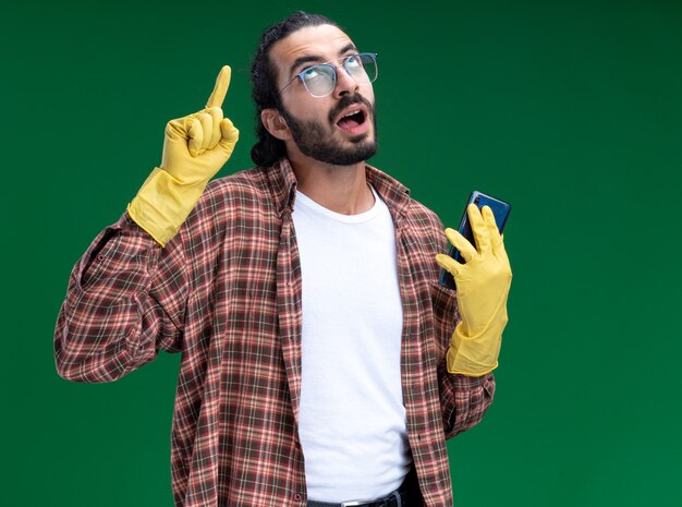 Impresionado joven guapo chico de limpieza con camiseta y guantes sosteniendo puntos de teléfono en aislado en la pared verde