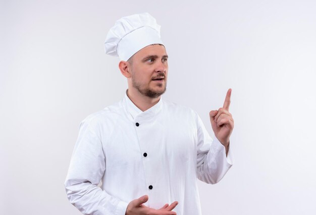 Impresionado joven guapo cocinero en uniforme de chef apuntando hacia arriba mirando al lado derecho aislado en la pared blanca