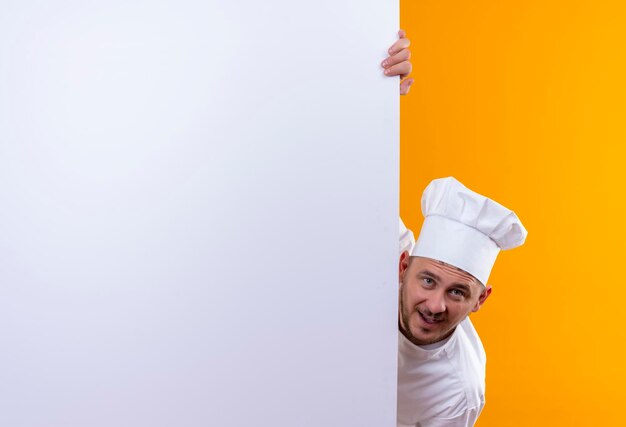 Impresionado joven guapo cocinero en uniforme de chef de pie detrás de una pared blanca y apuntando a ella aislada en la pared naranja con espacio de copia