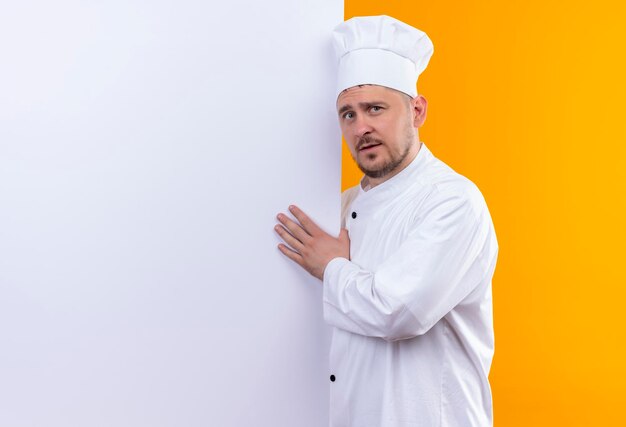 Impresionado joven guapo cocinero en uniforme de chef de pie detrás de la pared blanca apuntando con la mano a ella aislada en la pared naranja con espacio de copia