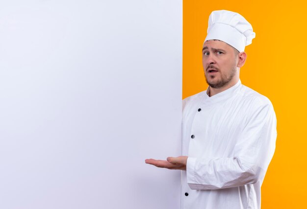 Impresionado joven guapo cocinero en uniforme de chef de pie detrás de la pared blanca apuntando con la mano a ella aislada en la pared naranja con espacio de copia