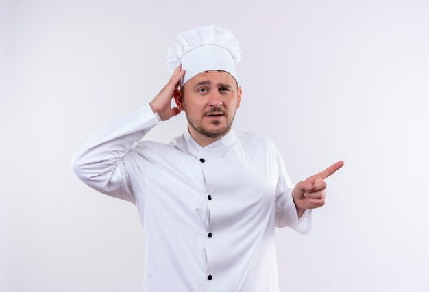 Impresionado joven guapo cocinero en uniforme de chef poniendo la mano en la cabeza y apuntando hacia el lado derecho aislado en la pared blanca