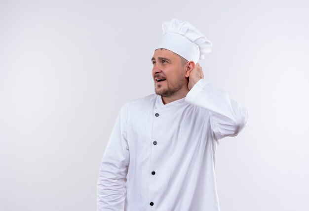 Impresionado joven guapo cocinero en uniforme de chef poniendo la mano detrás de la cabeza y mirando al lado aislado en la pared blanca