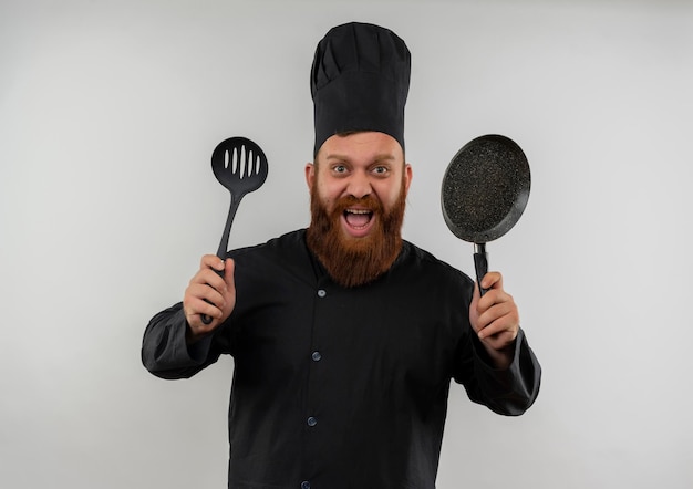 Impresionado joven guapo cocinero en uniforme de chef sosteniendo una cuchara ranurada y una sartén aislado en la pared blanca