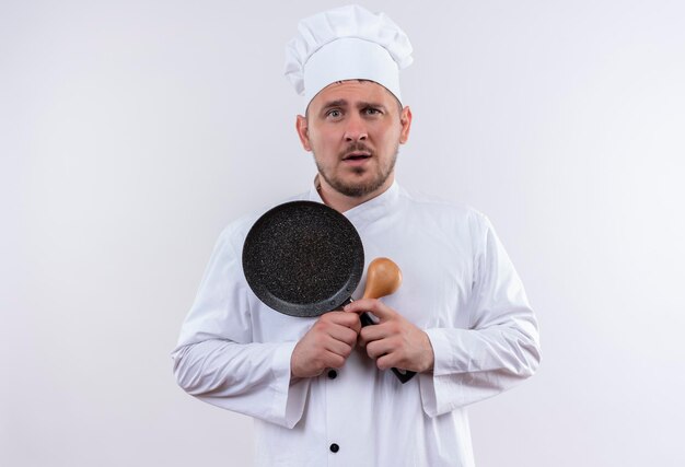 Impresionado joven guapo cocinero en uniforme de chef sosteniendo una cuchara y una sartén en la pared blanca aislada