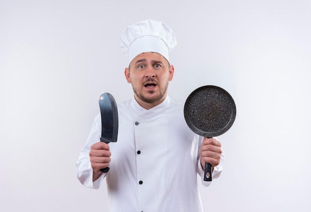 Impresionado joven guapo cocinero en uniforme de chef sosteniendo una cuchilla y una sartén en una pared blanca aislada con espacio de copia