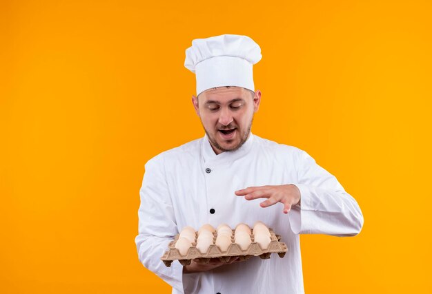 Impresionado joven guapo cocinero en uniforme de chef sosteniendo y mirando el cartón de huevos manteniendo la mano sobre él aislado en la pared naranja