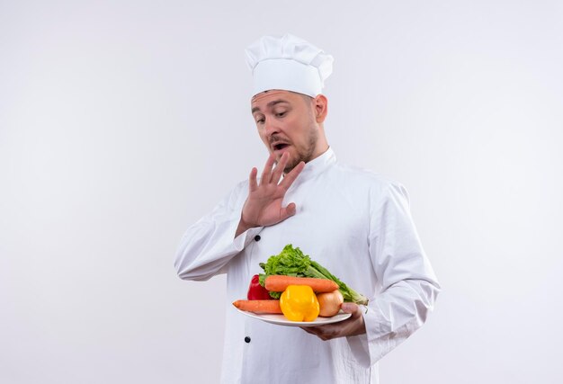 Impresionado joven guapo cocinero en uniforme de chef sosteniendo un plato con verduras y manteniendo la mano sobre ellos en una pared blanca aislada con espacio de copia