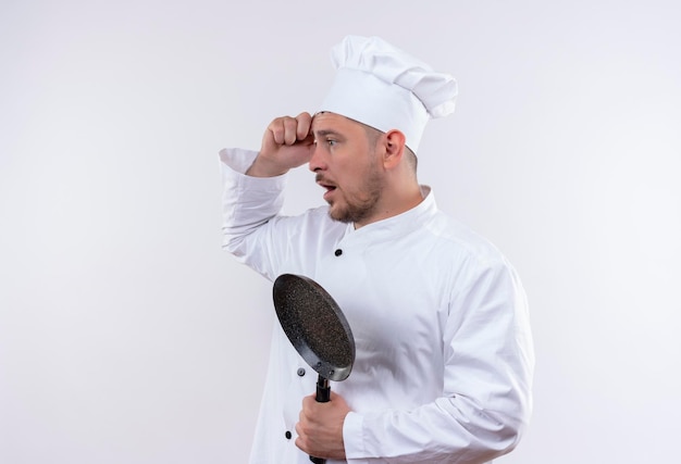 Impresionado joven guapo cocinero en uniforme de chef sosteniendo una sartén de pie en la vista de perfil mirando de lado con la mano en la cabeza aislada en la pared blanca