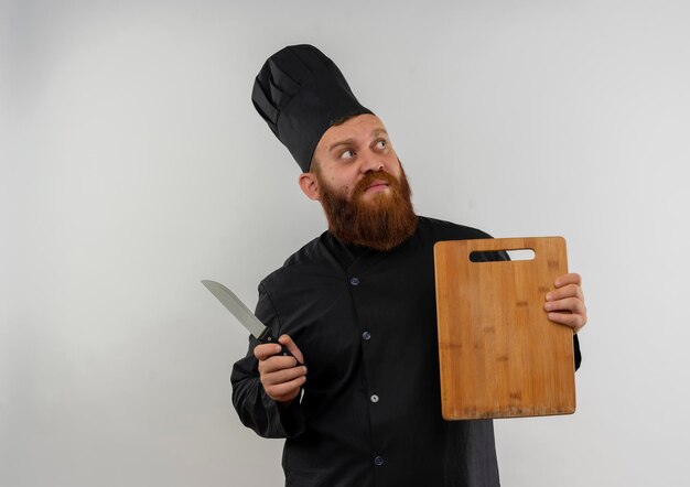 Impresionado joven guapo cocinero en uniforme de chef sosteniendo una tabla de cortar y un cuchillo mirando al lado aislado en la pared blanca con espacio de copia