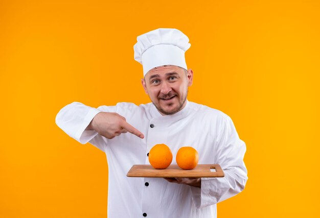 Impresionado joven guapo cocinero en uniforme de chef sosteniendo una tabla de cortar con naranjas apuntando a ellos aislados en la pared naranja