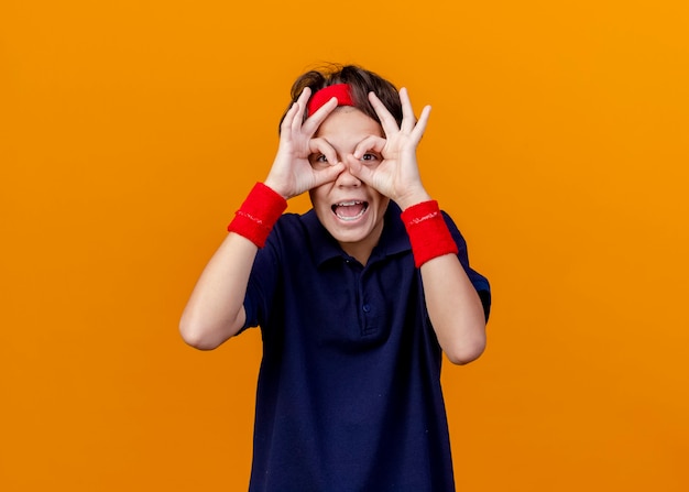 Impresionado joven guapo deportivo con diadema y muñequeras con aparatos dentales mirando a la cámara haciendo gestos de mirada con las manos como binoculares aislados sobre fondo naranja con espacio de copia