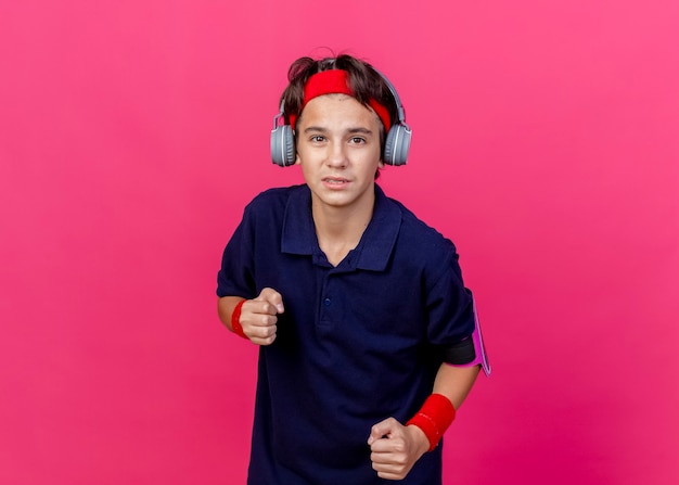 Impresionado joven guapo deportivo con diadema y muñequeras y auriculares brazalete de teléfono con aparatos dentales apretando los puños aislados en la pared carmesí con espacio de copia