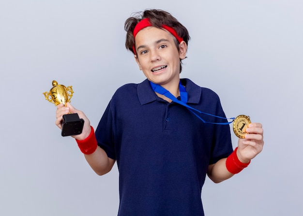 Impresionado joven guapo deportivo vistiendo diadema y muñequeras con aparatos dentales y medalla alrededor del cuello sosteniendo la medalla y la copa ganadora mirando a cámara aislada sobre fondo blanco