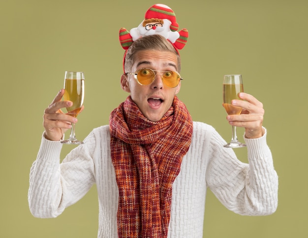 Foto gratuita impresionado joven guapo con diadema de santa claus y bufanda sosteniendo dos copas de champán aislado en la pared verde oliva