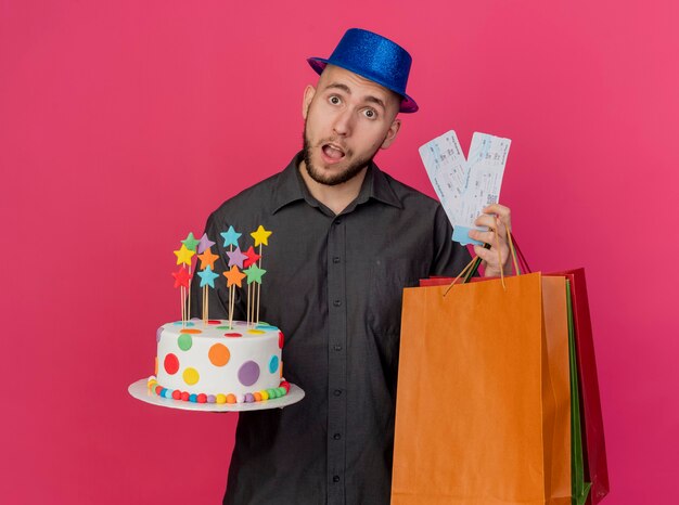 Foto gratuita impresionado joven guapo eslavo partido con sombrero de fiesta sosteniendo boletos de avión de pastel de cumpleaños y bolsas de papel mirando a cámara aislada sobre fondo carmesí con espacio de copia