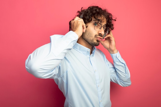 Impresionado joven guapo con gafas y auriculares mirando al frente tocando auriculares haciendo gesto de beso aislado en la pared rosa