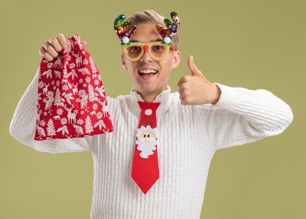 Impresionado joven guapo con gafas de Navidad y corbata de santa claus sosteniendo un saco de Navidad mostrando el pulgar hacia arriba aislado en la pared verde oliva