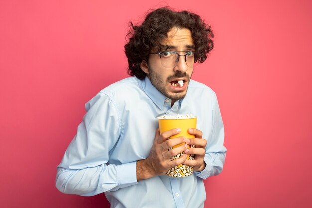 Impresionado joven guapo con gafas sosteniendo un cubo de palomitas de maíz mirando al frente con un trozo de palomitas de maíz en la boca aislada en la pared rosa