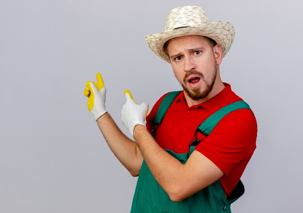 Impresionado joven guapo jardinero eslavo en uniforme con guantes de jardinería y sombrero mirando apuntando detrás aislado