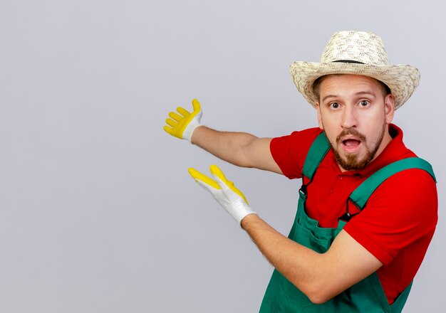 Impresionado joven guapo jardinero eslavo en uniforme con guantes de jardinería y sombrero mirando apuntando con las manos detrás de aislados