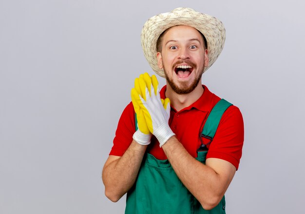 Impresionado joven guapo jardinero eslavo en uniforme con guantes de jardinería y sombrero mirando manteniendo las manos juntas aisladas