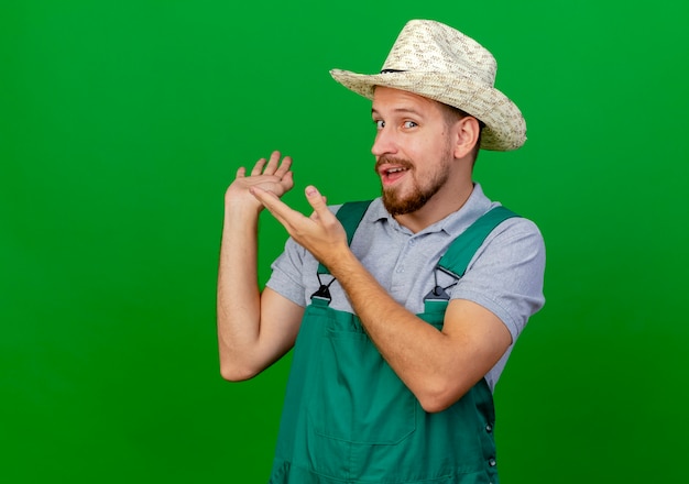 Foto gratuita impresionado joven guapo jardinero eslavo en uniforme y sombrero mirando apuntando con las manos detrás