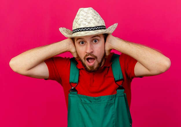 Impresionado joven guapo jardinero eslavo en uniforme y sombrero mirando poniendo las manos en las orejas