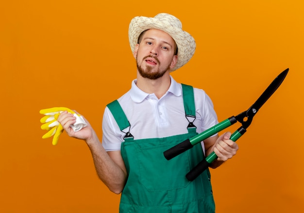 Impresionado joven guapo jardinero eslavo en uniforme con sombrero sosteniendo guantes de jardinería y podadoras mirando aislado en la pared naranja