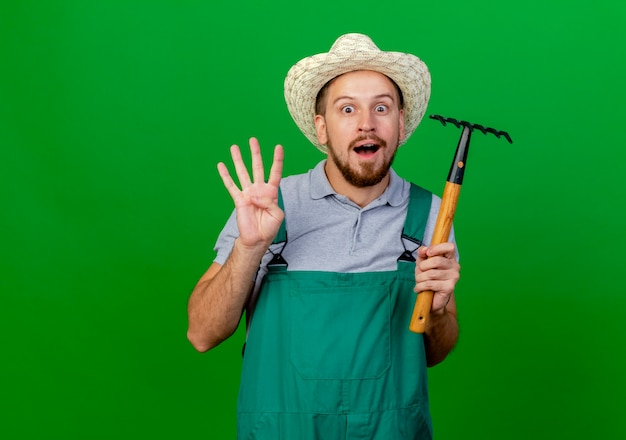 Foto gratuita impresionado joven guapo jardinero eslavo en uniforme y sombrero sosteniendo rastrillo mirando mostrando cuatro con la mano